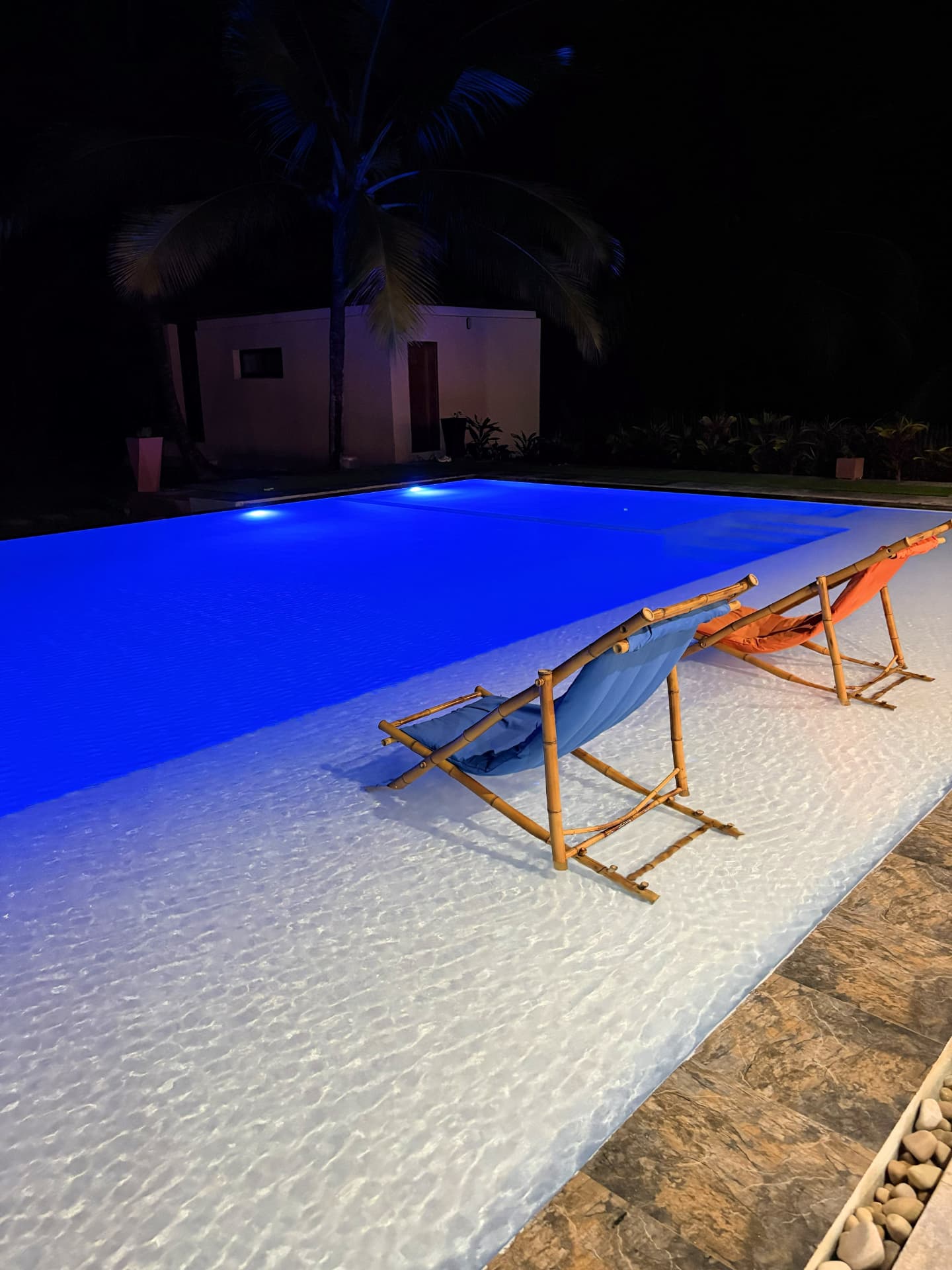 The pool lit blue after dark, deck chairs at the water's edge